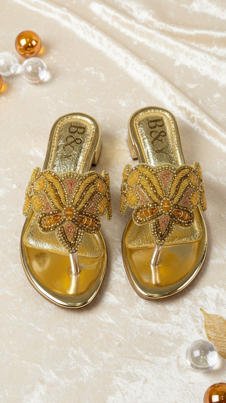 A pair of gold, beaded, and embellished sandals are displayed on a beige surface, surrounded by small decorative items.