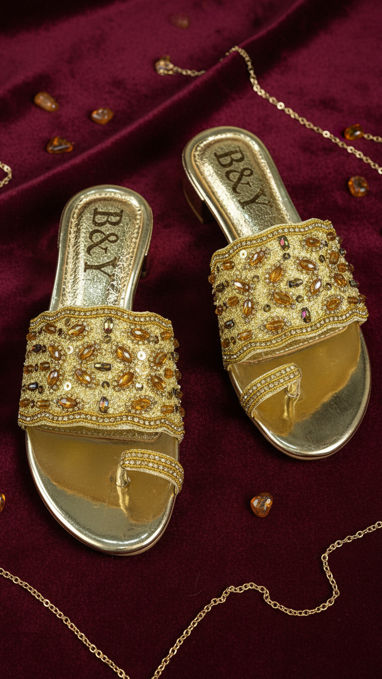 A pair of gold sandals with beaded embellishments, placed on a burgundy background with scattered small gold objects.