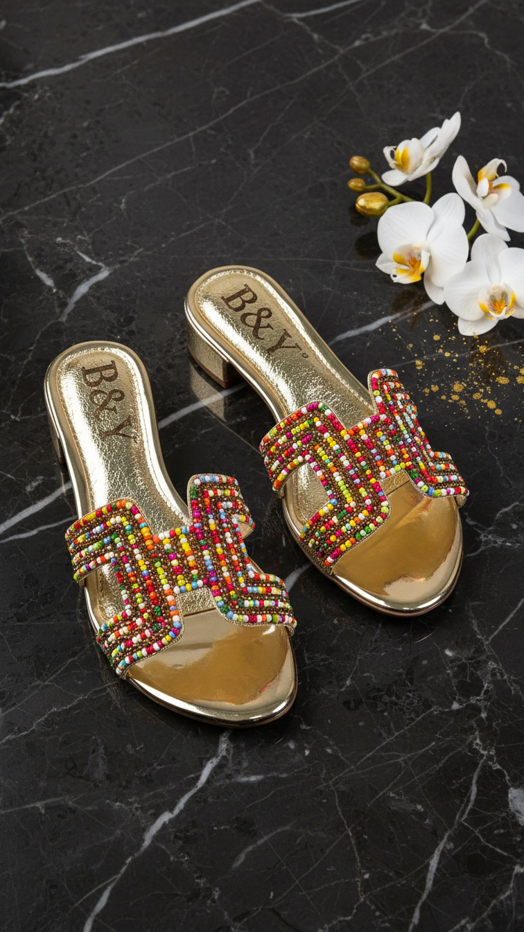 A pair of gold-colored, embellished sandals with colorful rhinestones are displayed on a black marble surface, accompanied by a small bouquet of white orchids.