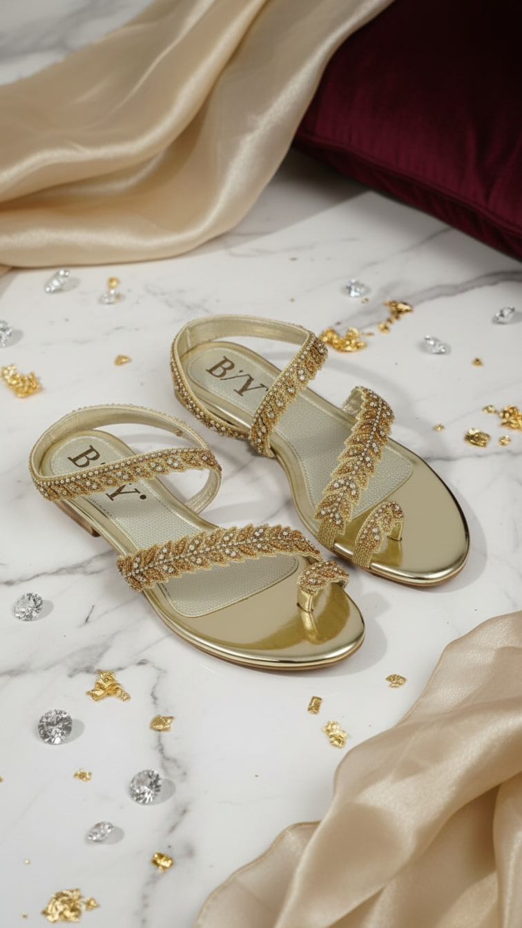 A pair of gold, beaded sandals with a leaf-like design, placed on a marble surface with scattered gold and silver glitter.