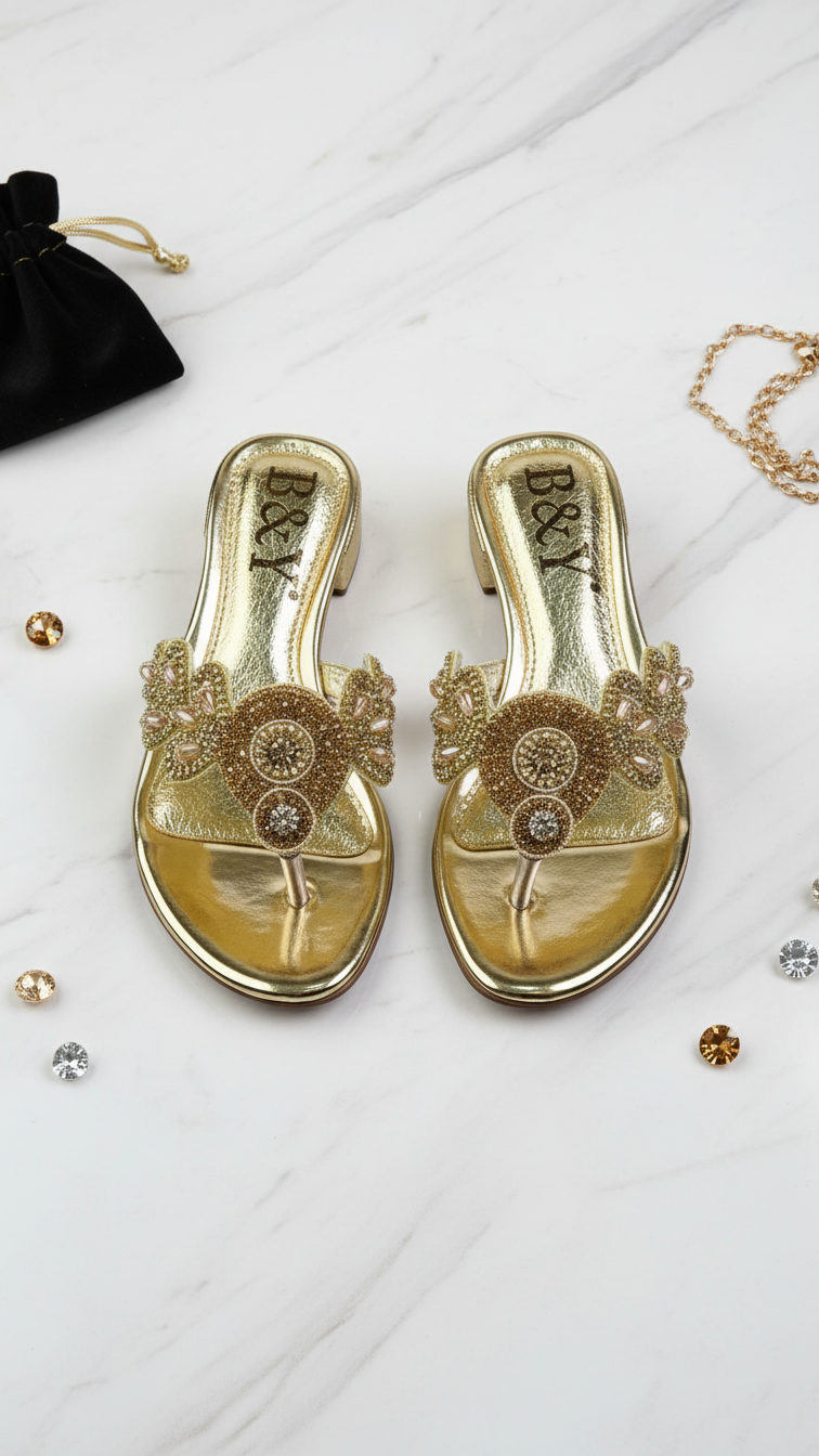 A pair of gold, embellished sandals with rhinestones and a chain are displayed on a white marble surface, accompanied by a small black bag and scattered gemstones.