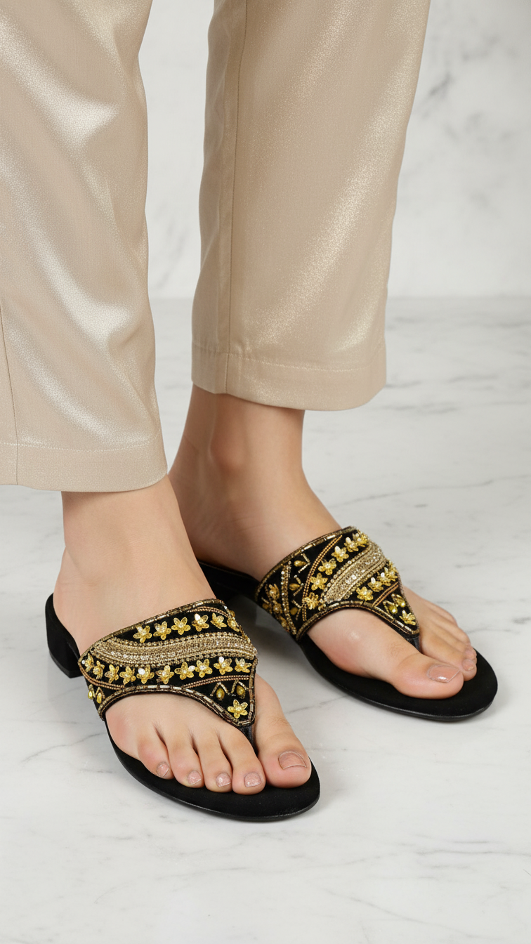 A pair of black and gold embellished sandals with gold accents, worn by a person standing on a marble floor.