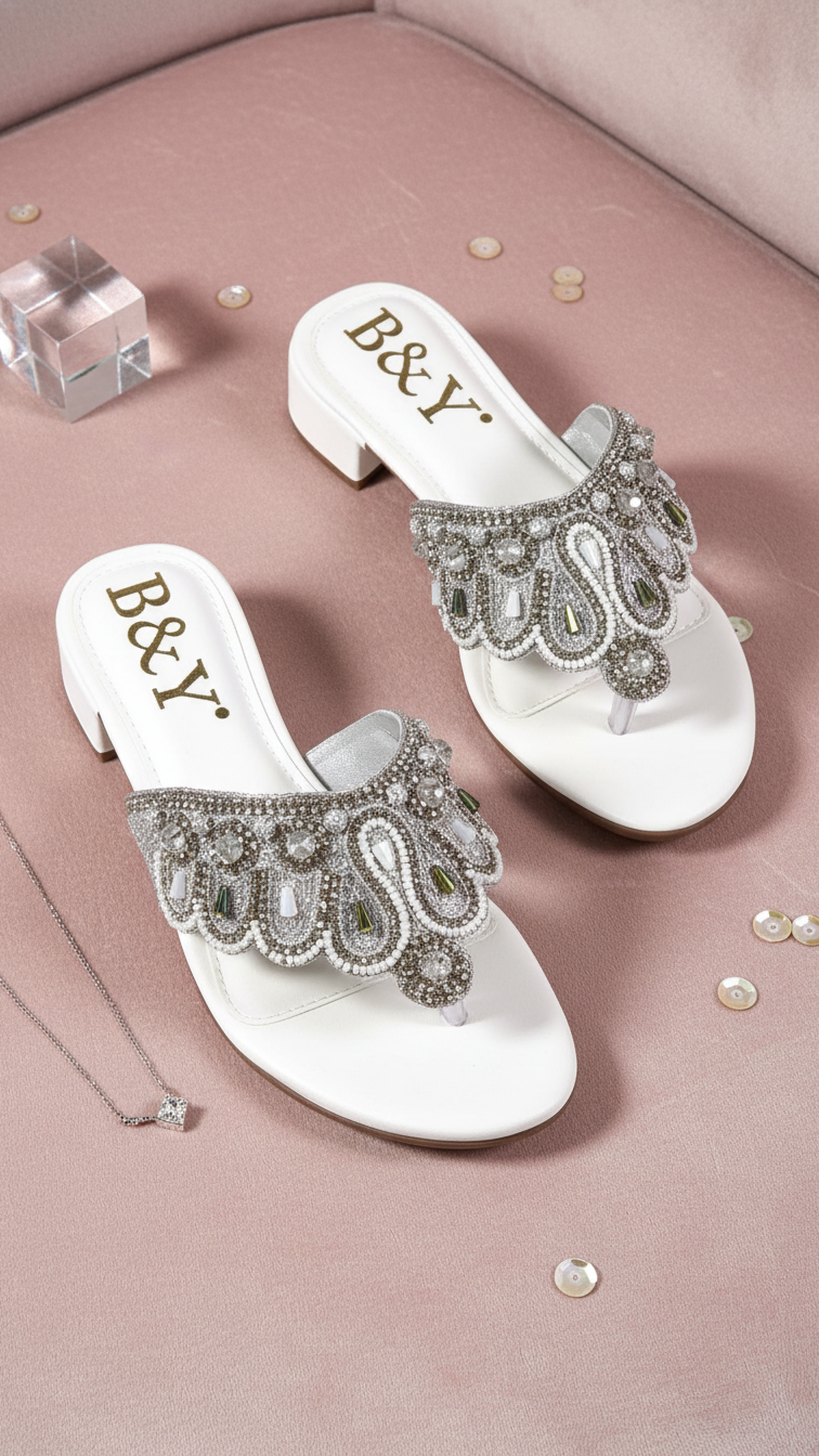 A pair of white, embellished sandals with rhinestones and a necklace are displayed on a pink surface.