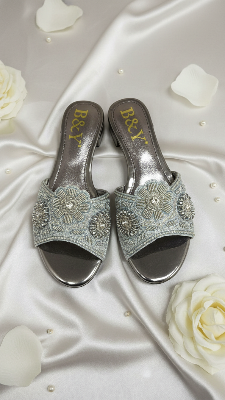 A pair of silver and blue embellished sandals with a floral design, placed on a white satin background with scattered white rose petals.