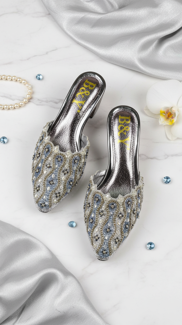 A pair of silver and blue embellished mens shoes with a gold logo, placed on a white marble surface alongside pearls, a white flower, and small blue gemstones.