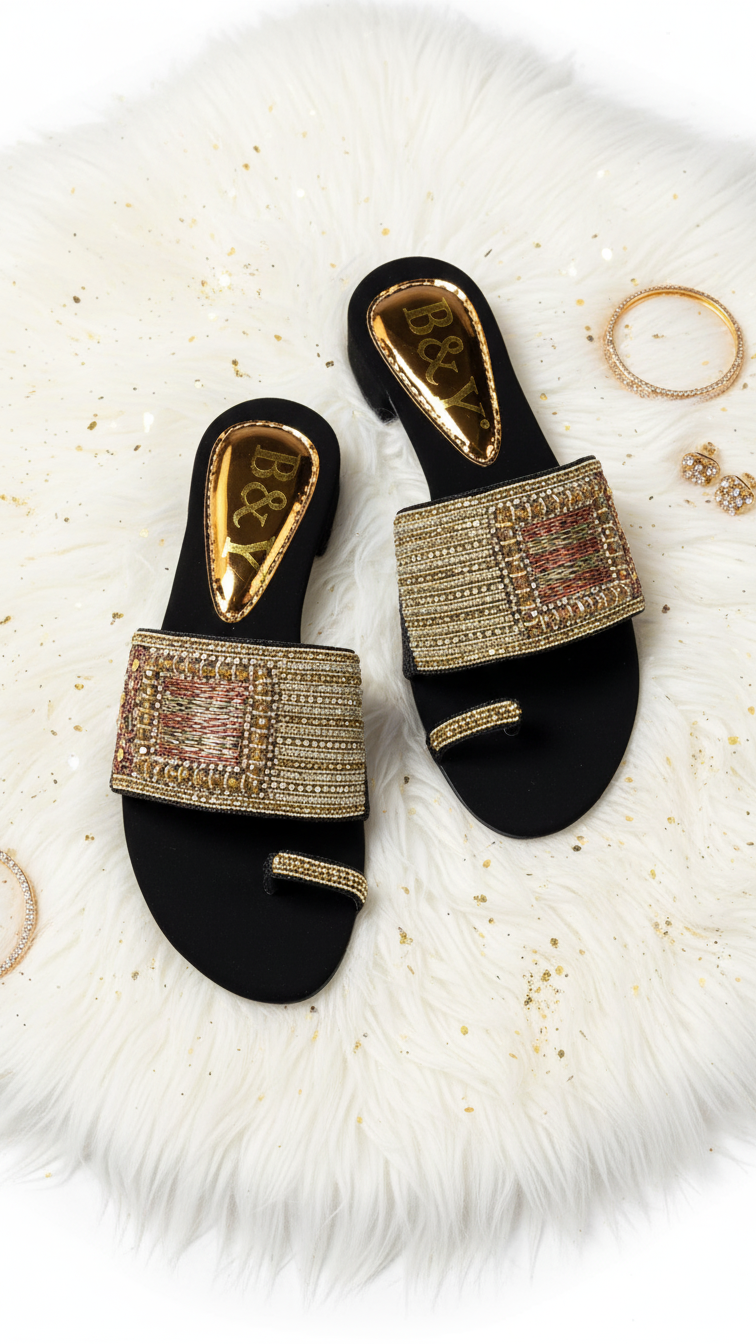 A pair of black sandals with gold embellishments, placed on a white fur rug with gold jewelry scattered around them.