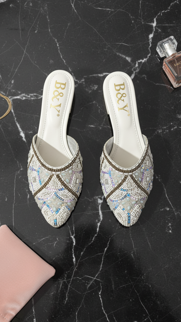 A pair of white, beaded, and embellished mens shoes with gold accents, placed on a black marble surface.