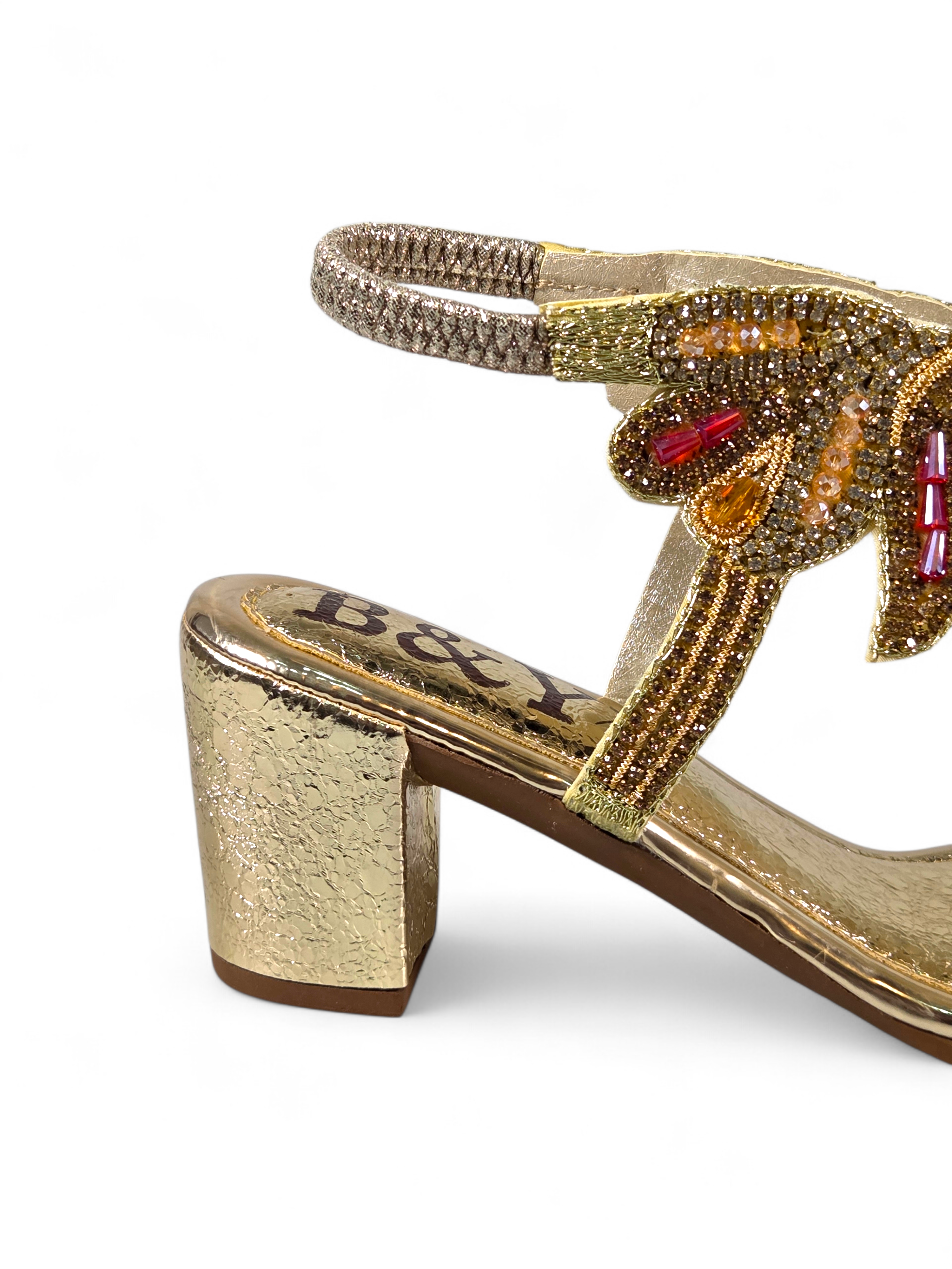 Sandália artesanal B&Y dourada com bordados manuais, cristais e pedrarias em tons de dourado e vermelho, salto médio e acabamento sofisticado, em fundo branco.