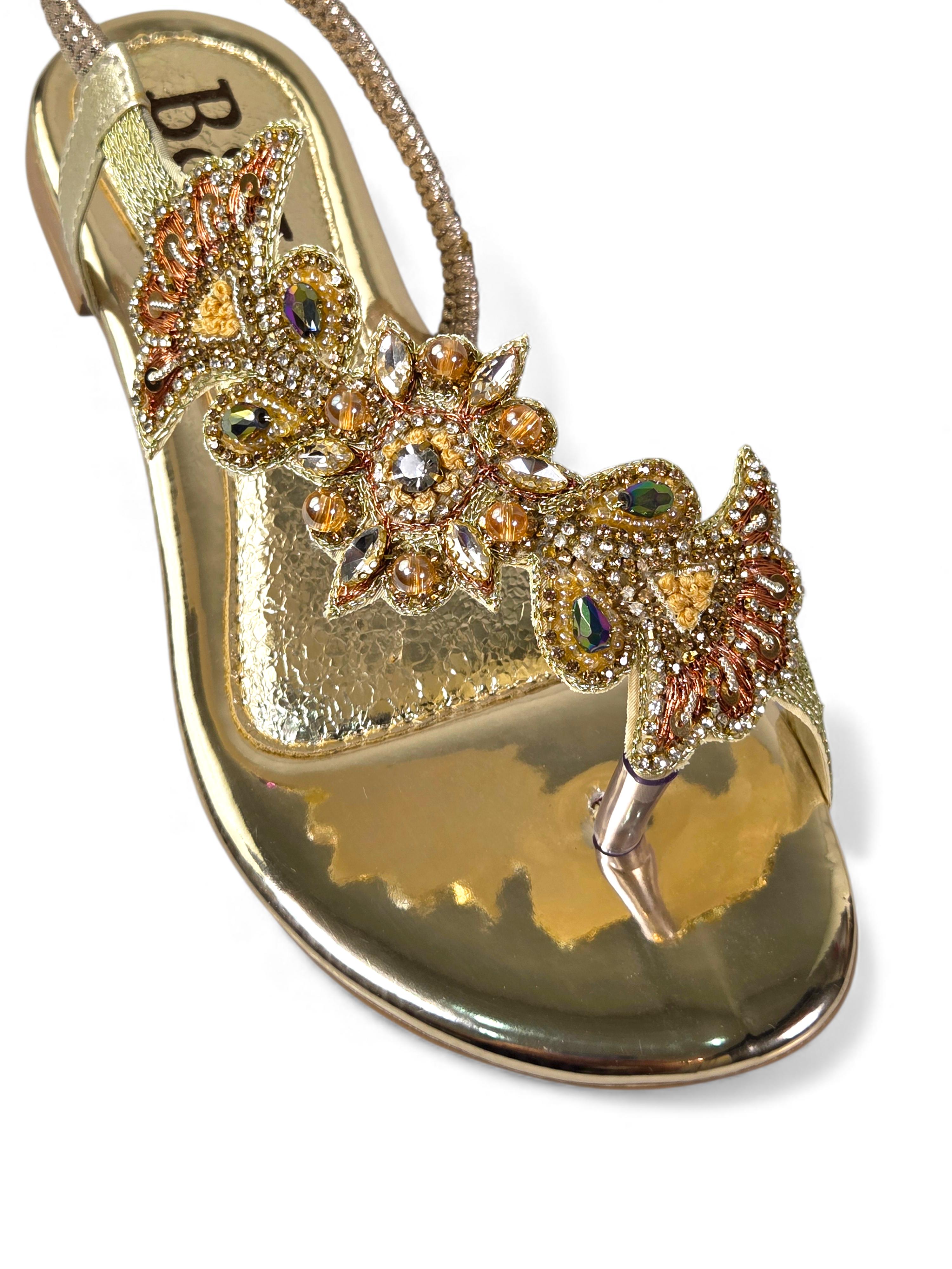 A close-up of a gold-colored sandal with intricate floral and gemstone embellishments.